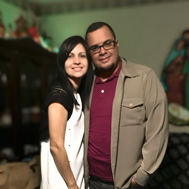 Israel Garcia-Diaz and his wife, both dark hair pose in a close hug and smile, facing the camera inside their home, blurred in the background.