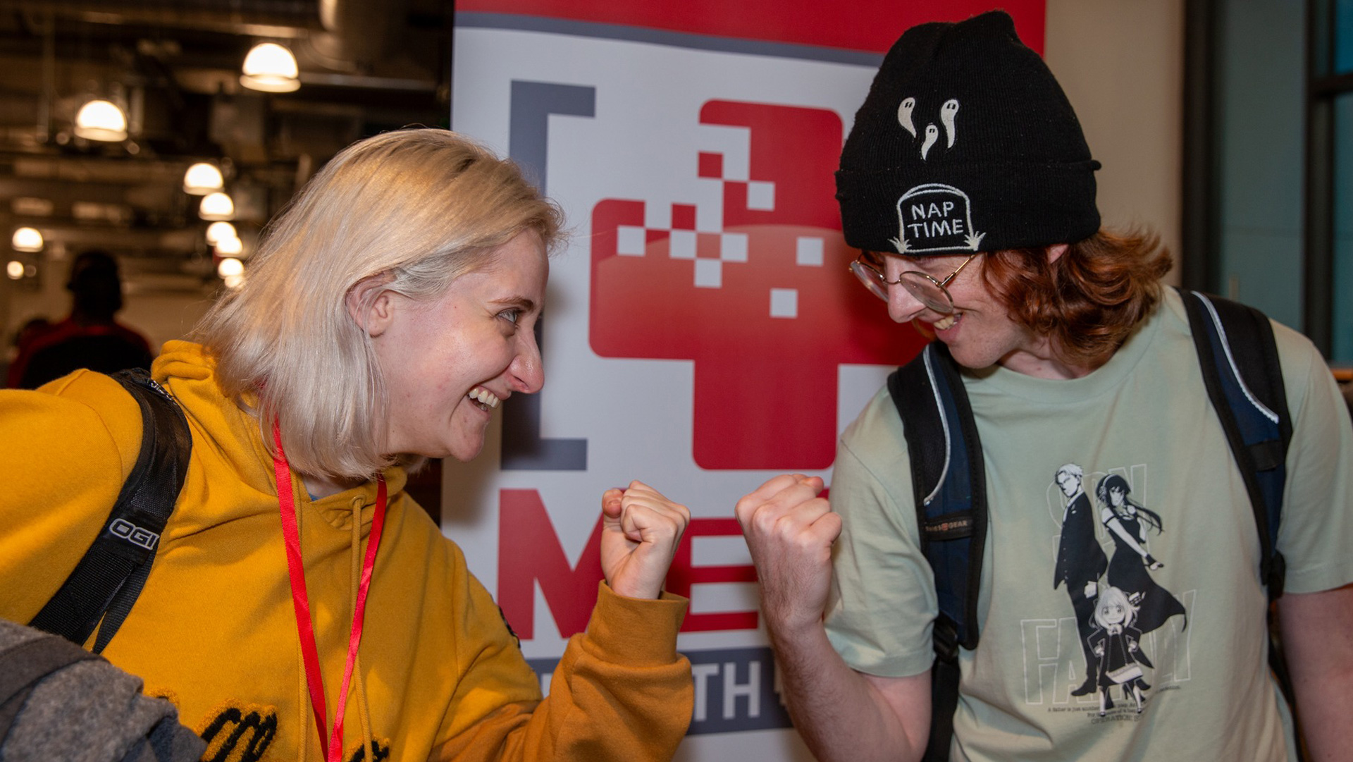 Two FIEA students stand facing each other with fists clenched in friendly competition wearing big smiles in front of the MeGa Health Jam logo.