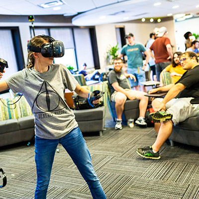 Young female student wearing VR gear playing in a large room with other students in the background