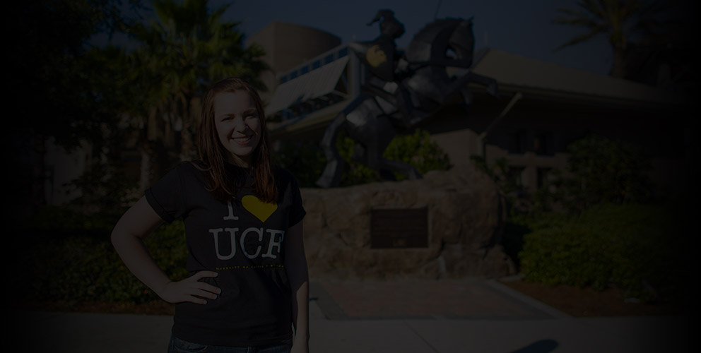 Woman with I heart UCF black tee on stands in front of a knight statue on a sunny day