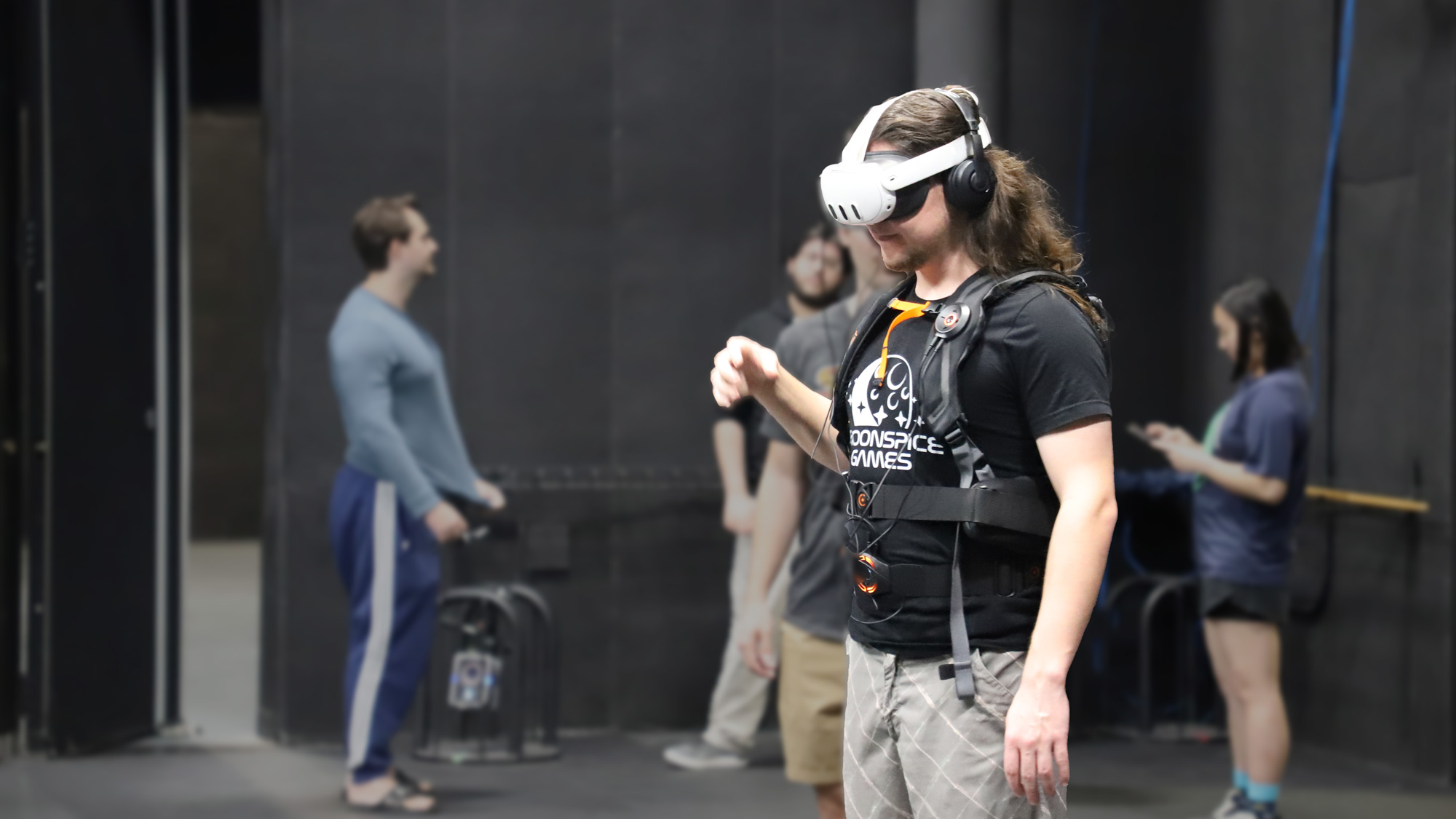 Cohort 21 students help each other suit up for the FIEA Funhouse experience in VR headset, noise cancelling headphones and rumble vest.