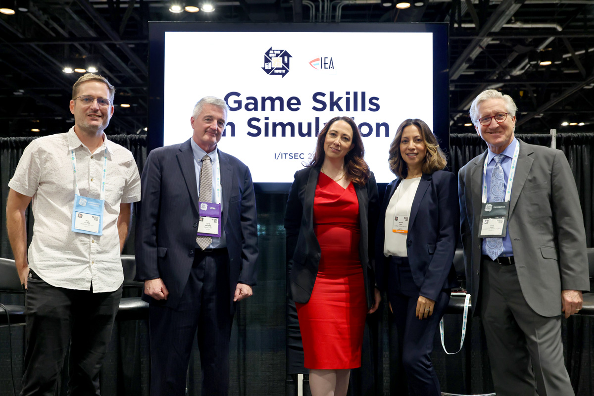 Four panelists and an instructor stand atop a black draped stage with a screen behind them that reads Game Skills in Simulation