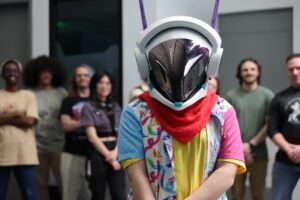 A person in a reflective helmet and colorful outfit stands in front of a group of people indoors.