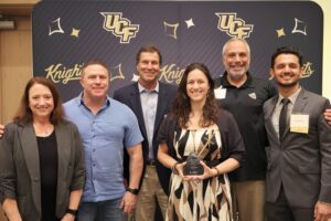 Six people pose for a group photo at a UCF Knights event; one woman in the center holds an award. UCF logos and "Knights" are visible on the backdrop behind them.
